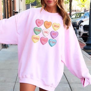 May include: Pink sweatshirt with a graphic of colorful candy hearts with phrases like "No Cap", "Bruh", "Sigma", "Ohio", "Rizzz", "Skibidi", "Toilet", and "V Day".