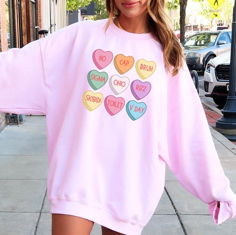 May include: Pink sweatshirt with a graphic of colorful candy hearts with phrases like "No Cap", "Bruh", "Sigma", "Ohio", "Rizzz", "Skibidi", "Toilet", and "V Day".