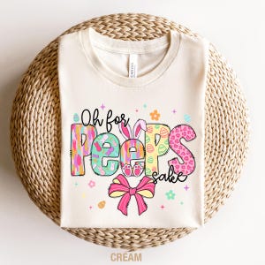 May include: A cream colored t-shirt with the text "Oh for Peep's sake" in colorful letters with a pink bow and Easter egg designs.