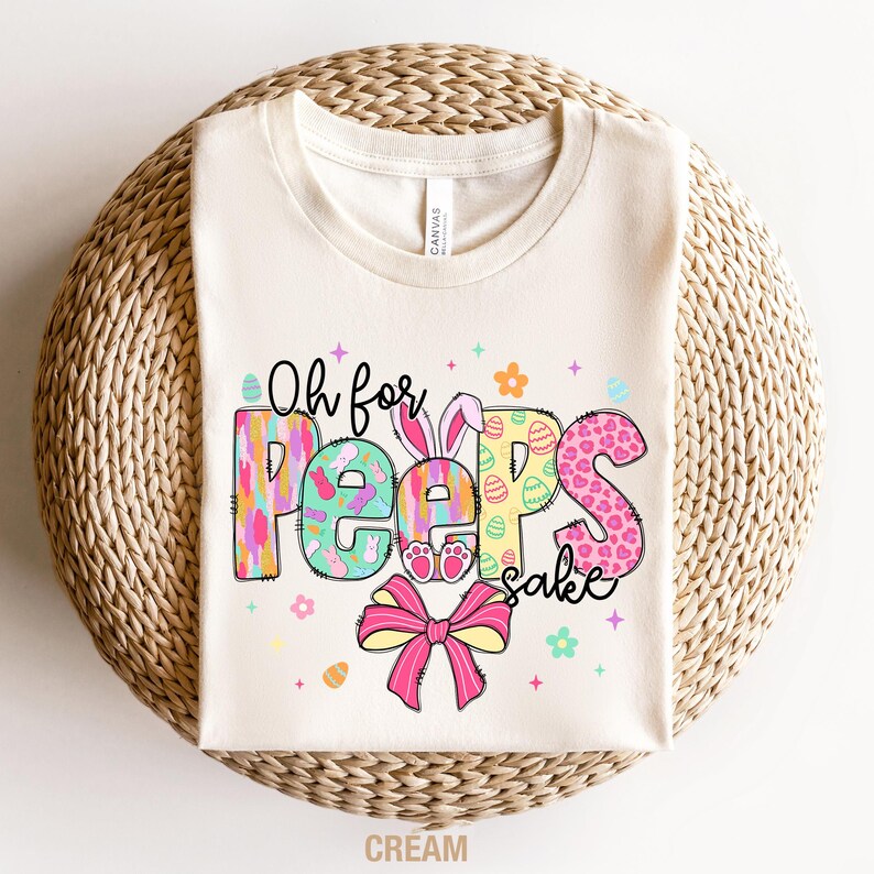 May include: A cream colored t-shirt with the text "Oh for Peep's sake" in colorful letters with a pink bow and Easter egg designs.