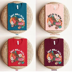 May include: Four t-shirts in teal, heather peach, red, and maroon colors. Each shirt features a graphic of a pink roller skate with white daisies, a rainbow arc, and the text "JUST ROLL WITH IT" printed in a white, curved font.