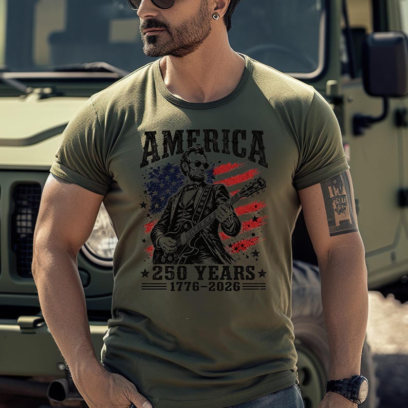 K&ouml;nnte beinhalten: Olivgr&uuml;nes T-Shirt mit dem Wort "AMERICA" &uuml;ber einer Grafik einer Person, die Gitarre vor einer rot-wei&szlig;-blauen Flagge spielt. Der Text "250 YEARS 1776-2026" befindet sich darunter.