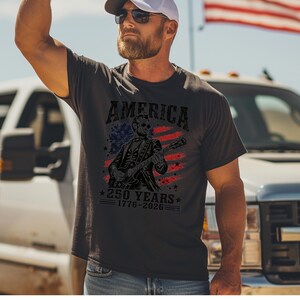 K&ouml;nnte beinhalten: Dunkelgraues T-Shirt mit dem Wort "AMERICA" &uuml;ber einer Illustration einer Figur, die Gitarre spielt, vor dem Hintergrund der amerikanischen Flagge. Der Text "250 YEARS 1776-2026" steht darunter.