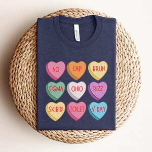 May include: A navy blue t-shirt with a design of nine candy hearts. Each heart has a different color and a word printed on it. The words are: NO, CAP, BRUH, SIGMA, OHIO, RIZZ, SKIBIDI, TOILET, and V DAY.