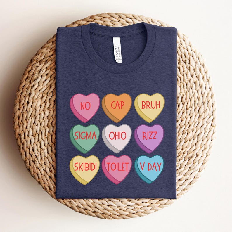 May include: A navy blue t-shirt with a design of nine candy hearts. Each heart has a different color and a word printed on it. The words are: NO, CAP, BRUH, SIGMA, OHIO, RIZZ, SKIBIDI, TOILET, and V DAY.