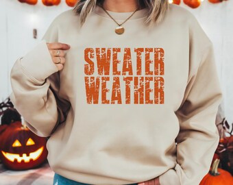 May include: Beige sweatshirt with the text "SWEATER WEATHER" in orange, distressed lettering. The sweatshirt is worn by a person. The background includes fall decorations like pumpkins and string lights.