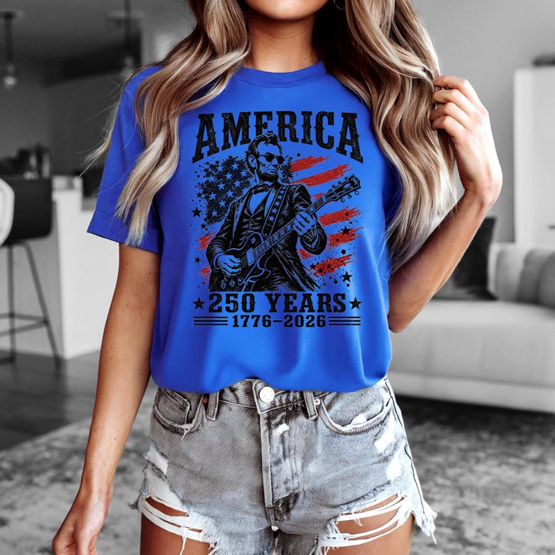 K&ouml;nnte beinhalten: K&ouml;nigsblaues T-Shirt mit einer Grafik, die das Wort "AMERICA" &uuml;ber einer Illustration von Abraham Lincoln zeigt, der Gitarre spielt. Das Design enth&auml;lt eine Flagge und den Text "250 YEARS 1776-2026."