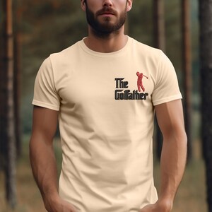 May include: A beige t-shirt with a black and red graphic that says "The Golfather" with a golfer silhouette.