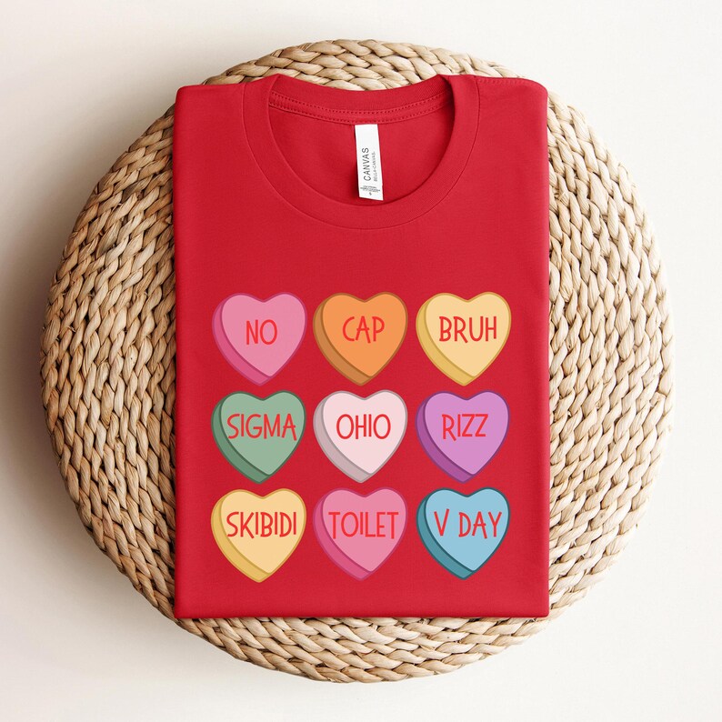 May include: Red t-shirt with nine colorful candy hearts printed on the front. Each heart has a different slang word printed on it, including "No", "Cap", "Bruh", "Sigma", "Ohio", "Rizz", "Skibidi", "Toilet", and "V Day".