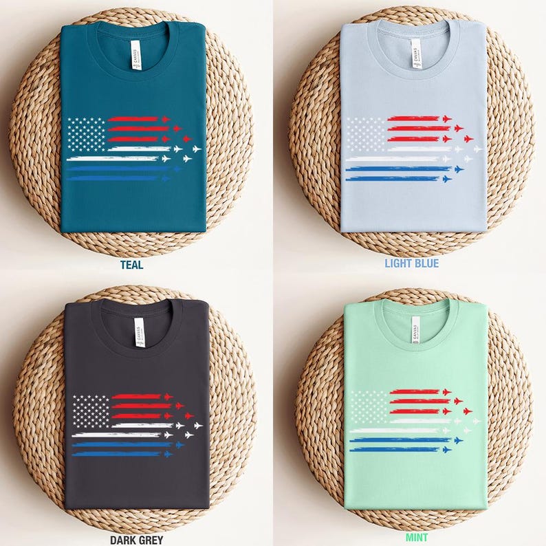 May include: Four t-shirts in teal, light blue, dark grey, and mint green, each featuring a graphic of the American flag with red and white stripes, a blue stripe, and a formation of white jets. The shirts are displayed on woven mats.