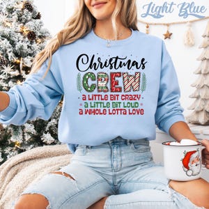 May include: Light blue Christmas Crew sweatshirt with the text "Christmas Crew" in black and red lettering. The sweatshirt also has the phrases "a little bit crazy", "a little bit loud", and "a whole lotta love". A white mug with a Santa Claus graphic is visible.