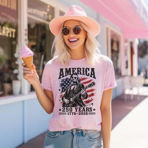 K&ouml;nnte beinhalten: Hellrosa T-Shirt mit dem Wort "AMERICA" in fetten schwarzen Buchstaben. Das Shirt zeigt eine Illustration von Abraham Lincoln, der Gitarre spielt, vor dem Hintergrund der amerikanischen Flagge. Der Text "250 YEARS 1776-2026" steht darunter.