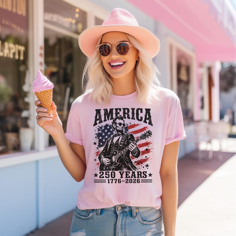 K&ouml;nnte beinhalten: Hellrosa T-Shirt mit dem Wort "AMERICA" in fetten schwarzen Buchstaben. Das Shirt zeigt eine Illustration von Abraham Lincoln, der Gitarre spielt, vor dem Hintergrund der amerikanischen Flagge. Der Text "250 YEARS 1776-2026" steht darunter.