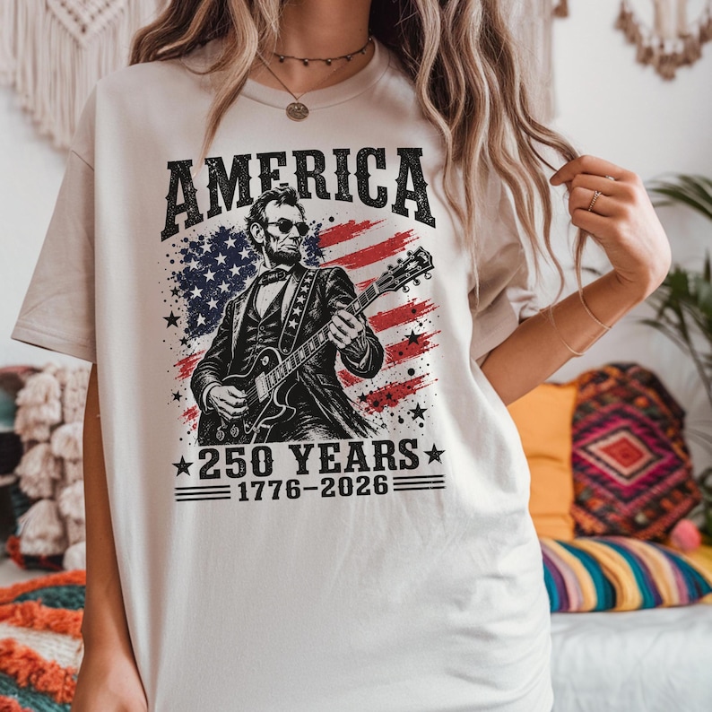 K&ouml;nnte beinhalten: Cremefarbenes T-Shirt mit einer Grafik von Abraham Lincoln, der Gitarre spielt, vor dem Hintergrund der amerikanischen Flagge. Der Text "AMERICA" steht &uuml;ber dem Bild, darunter "250 YEARS 1776-2026". Ein patriotisches Design.