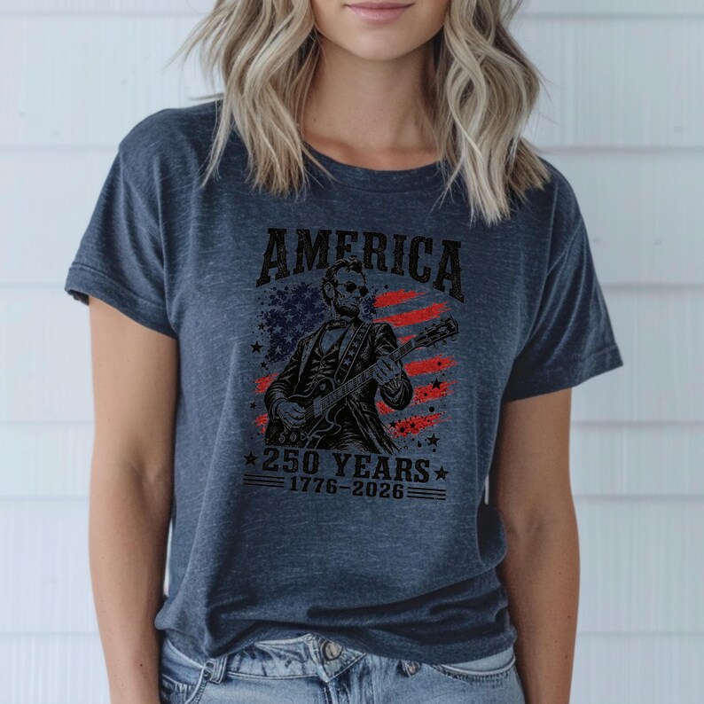 K&ouml;nnte beinhalten: Marineblaues T-Shirt mit dem Wort "AMERICA" &uuml;ber einer Illustration einer Figur, die Gitarre vor einem US-Flaggen-Design spielt. Der Text "250 YEARS 1776-2026" befindet sich darunter.