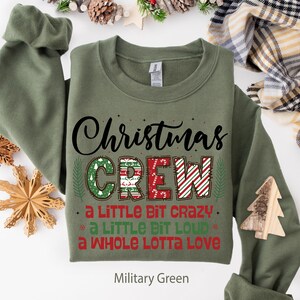 May include: Military green sweatshirt with the text "Christmas CREW" in a festive design. The word "CREW" is decorated with Christmas patterns and the phrases "a LITTLE BIT CRAZY", "a LITTLE BIT LOUD", and "a WHOLE LOTTA LOVE".