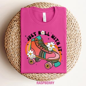 May include: A raspberry pink t-shirt featuring a graphic of a coral and teal roller skate surrounded by white daisies. The text "JUST ROLL WITH IT" is printed in black above the skate, which is set against a circular orange and yellow rainbow.