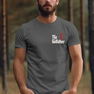 May include: A dark grey t-shirt with a white graphic that says "The Golfather" with a red golfer silhouette.