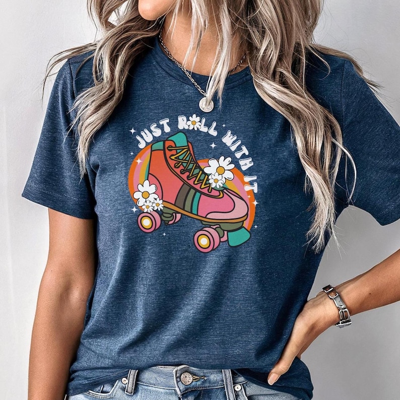 May include: A dark blue heathered t-shirt featuring a graphic of a pink and orange roller skate surrounded by white daisies and a rainbow arc. The text "JUST ROLL WITH IT" is printed in white letters above the roller skate illustration.