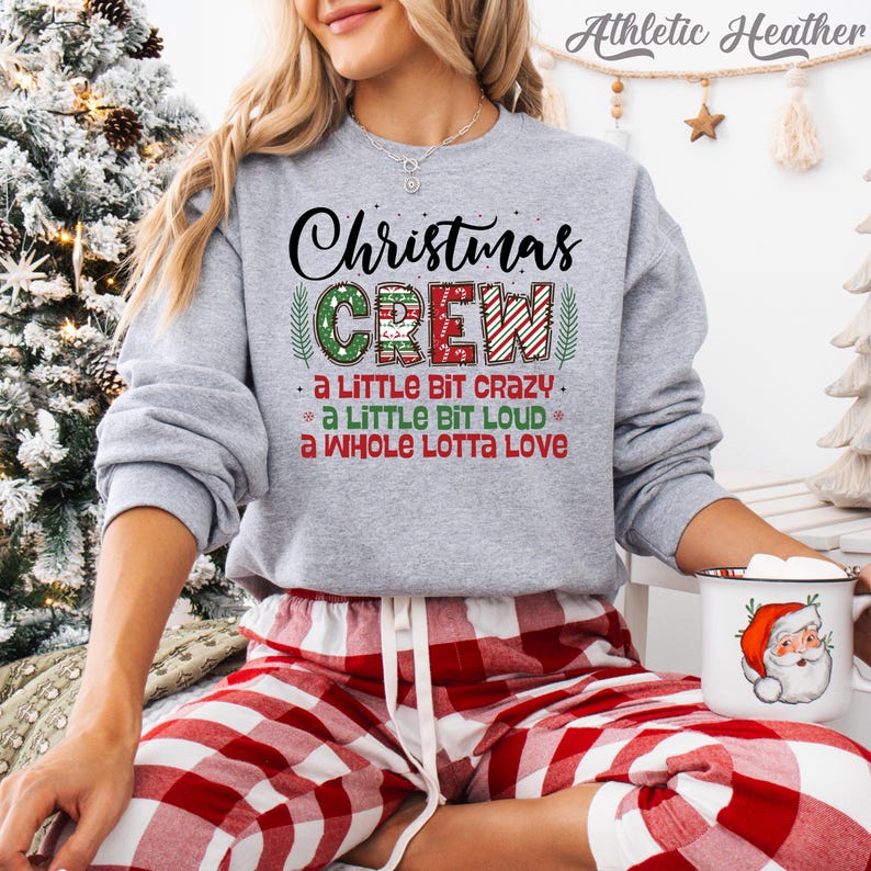 May include: A gray sweatshirt with the text "Christmas CREW" in black, green, and red. The sweatshirt also has the phrases "A LITTLE BIT CRAZY," "A LITTLE BIT LOUD," and "A WHOLE LOTTA LOVE." The person is wearing red and white plaid pajama pants and holding a mug with a Santa Claus design.
