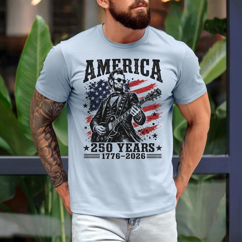 K&ouml;nnte beinhalten: Hellblaues T-Shirt mit einer Grafik von Abraham Lincoln, der Gitarre spielt, vor einer amerikanischen Flagge. Der Text auf dem Shirt lautet "AMERICA" und "250 YEARS 1776-2026".