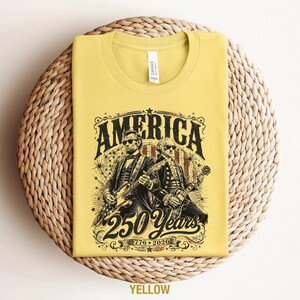 May include: Yellow t-shirt with a graphic featuring Abraham Lincoln and George Washington playing guitars, with the word "AMERICA" above and "250 Years 1776-2026" below. The shirt is folded on a woven surface.