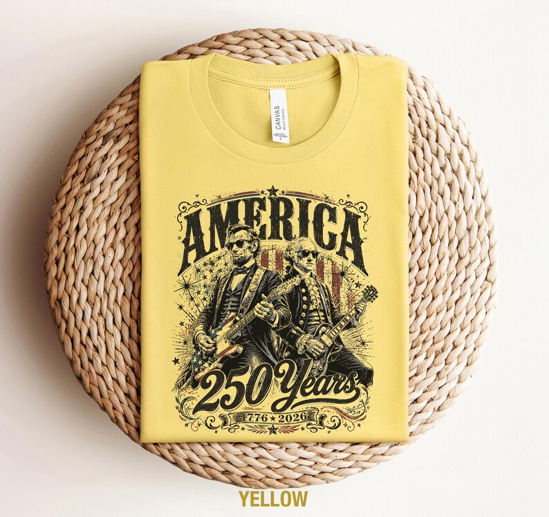 May include: Yellow t-shirt with a graphic featuring Abraham Lincoln and George Washington playing guitars, with the word "AMERICA" above and "250 Years 1776-2026" below. The shirt is folded on a woven surface.