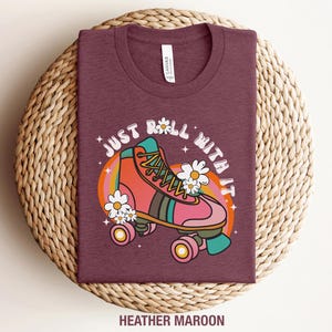 May include: A heather maroon t-shirt featuring a graphic of a coral and teal roller skate surrounded by white daisies and a rainbow arc. The text "JUST ROLL WITH IT" is printed in a white curved font above the skate, with small stars.