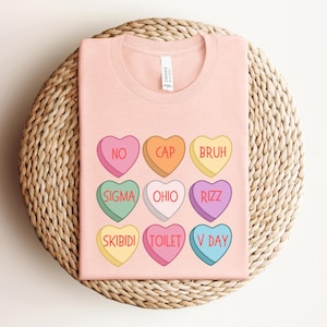 May include: Pink t-shirt with a design of nine candy hearts with different phrases on them. The phrases are "No", "Cap", "Bruh", "Sigma", "Ohio", "Rizz", "Skibidi", "Toilet", and "V Day".