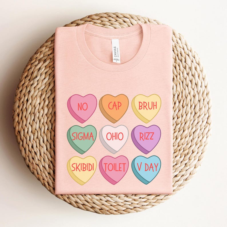 May include: Pink t-shirt with a design of nine candy hearts with different phrases on them. The phrases are "No", "Cap", "Bruh", "Sigma", "Ohio", "Rizz", "Skibidi", "Toilet", and "V Day".