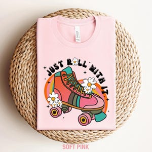 May include: A soft pink t-shirt featuring a graphic of a pink and orange roller skate surrounded by white daisies. The text "JUST ROLL WITH IT" is printed in black above the skate, which is set against a background of orange and yellow arches.
