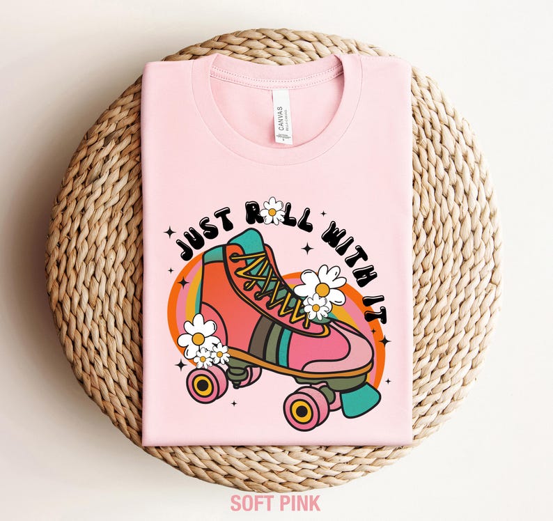 May include: A soft pink t-shirt featuring a graphic of a pink and orange roller skate surrounded by white daisies. The text "JUST ROLL WITH IT" is printed in black above the skate, which is set against a background of orange and yellow arches.