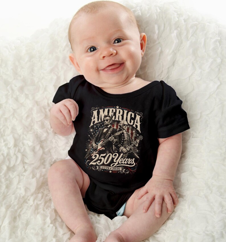 May include: A smiling baby wearing a black onesie with the text "AMERICA 250 Years 1776-2026" and an illustration of two figures playing guitars. The baby is lying on a white, textured surface.