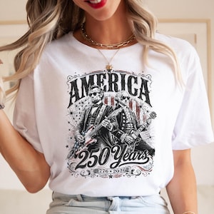 May include: White t-shirt featuring a graphic design with the word "AMERICA" above an illustration of two figures playing guitars. The design includes the text "250 Years" and the dates "1776-2026".