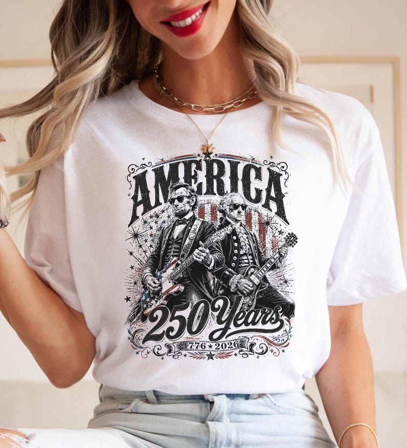 May include: White t-shirt featuring a graphic design with the word "AMERICA" above an illustration of two figures playing guitars. The design includes the text "250 Years" and the dates "1776-2026".