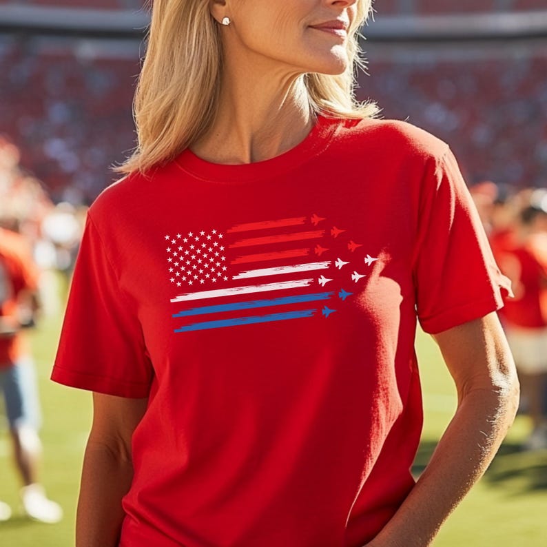 May include: Red t-shirt featuring a stylized American flag design. The flag incorporates white stars, red and white stripes, and blue stripes with jet plane silhouettes. The shirt is short-sleeved.