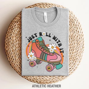 May include: A heather gray t-shirt featuring a graphic of a pink roller skate with teal and brown accents, surrounded by white daisies and the text "JUST ROLL WITH IT" in a black arched font. The design includes a colorful orange and yellow sunburst.
