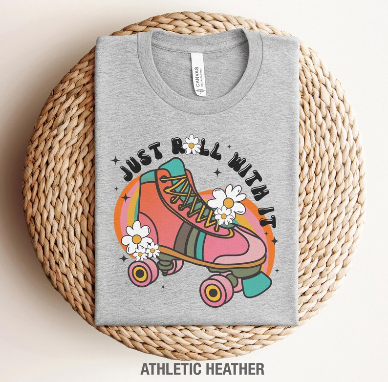 May include: A heather gray t-shirt featuring a graphic of a pink roller skate with teal and brown accents, surrounded by white daisies and the text "JUST ROLL WITH IT" in a black arched font. The design includes a colorful orange and yellow sunburst.