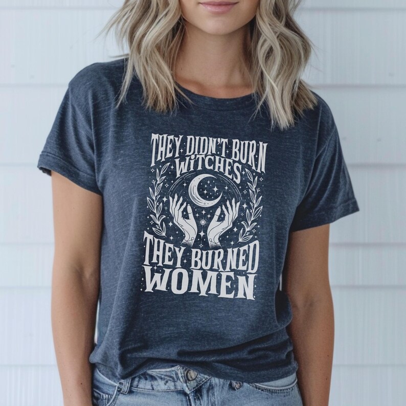 May include: Dark blue t-shirt featuring the white text "THEY DIDN'T BURN WITCHES THEY BURNED WOMEN". The design includes a crescent moon, stars, and hands. The shirt has a crew neck and short sleeves.
