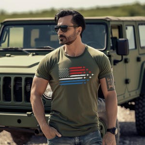 May include: Olive green t-shirt featuring a stylized American flag design. The flag incorporates red, white, and blue stripes with a formation of white airplanes. The shirt is short-sleeved and has a crew neck.