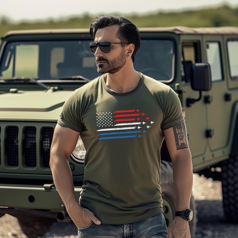 May include: Olive green t-shirt featuring a stylized American flag design. The flag incorporates red, white, and blue stripes with a formation of white airplanes. The shirt is short-sleeved and has a crew neck.