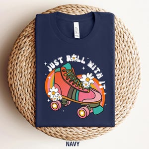 May include: A navy blue t-shirt featuring a graphic of a pink roller skate with green and orange accents, surrounded by white daisies and a rainbow arc. The text "JUST ROLL WITH IT" is printed in a white, curved, bold, retro-style font above.