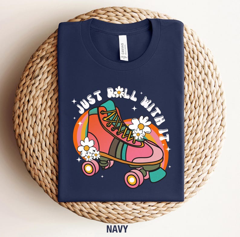 May include: A navy blue t-shirt featuring a graphic of a pink roller skate with green and orange accents, surrounded by white daisies and a rainbow arc. The text "JUST ROLL WITH IT" is printed in a white, curved, bold, retro-style font above.