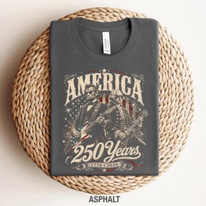 May include: Dark gray t-shirt with a graphic featuring Abraham Lincoln and George Washington playing guitars. The design includes the word "AMERICA" and "250 Years 1776-2026" in a vintage style. The shirt is made of a soft material.