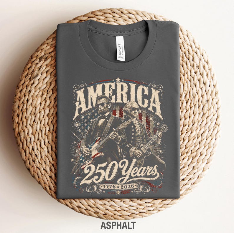 May include: Dark gray t-shirt with a graphic featuring Abraham Lincoln and George Washington playing guitars. The design includes the word "AMERICA" and "250 Years 1776-2026" in a vintage style. The shirt is made of a soft material.