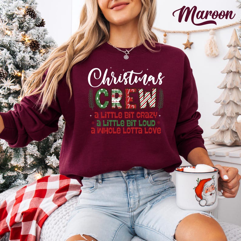 May include: Maroon Christmas Crew sweatshirt with white text. The design includes the words "Christmas Crew" and the phrases "a little bit crazy," "a little bit loud," and "a whole lotta love." The sweatshirt is a festive holiday apparel item.