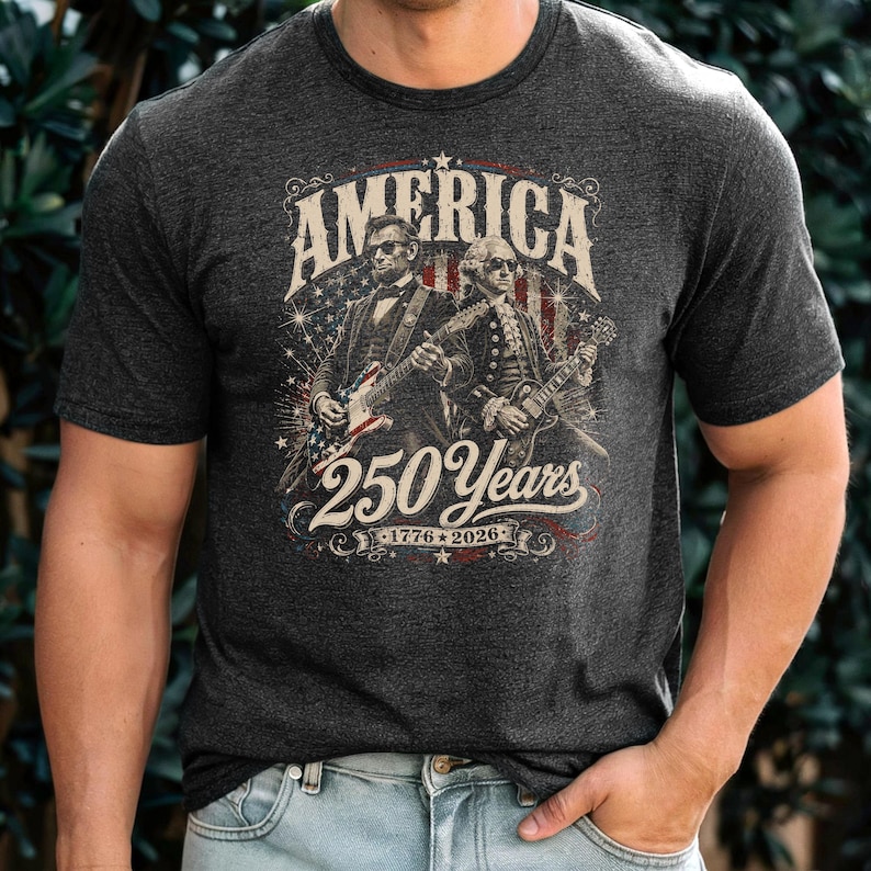 May include: Dark gray t-shirt featuring a graphic with the word "AMERICA" above an illustration of two historical figures playing electric guitars. The graphic also includes the text "250 Years 1776-2026" in a patriotic design.