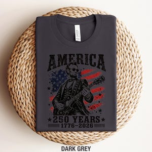 K&ouml;nnte beinhalten: Dunkelgraues T-Shirt mit einer Grafik von Abraham Lincoln, der Gitarre spielt, vor einer amerikanischen Flagge. Das Wort "AMERICA" steht &uuml;ber dem Bild, darunter "250 YEARS 1776-2026". Das T-Shirt liegt gefaltet auf einer gewebten Oberfl&auml;che.