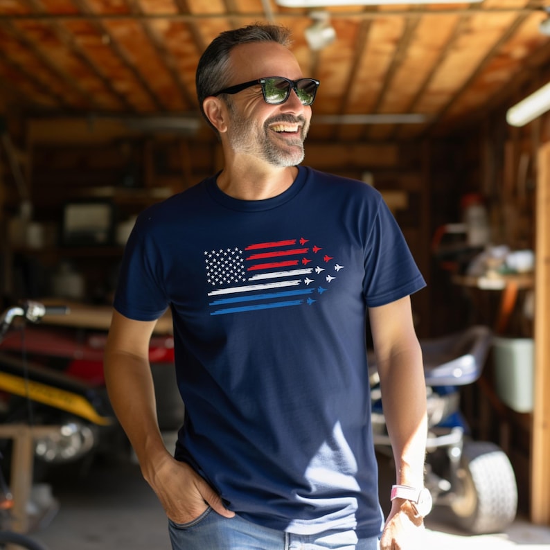 May include: Navy blue t-shirt featuring a stylized American flag design. The flag incorporates the stars and stripes with red, white, and blue accents, and includes a formation of jet aircraft. The shirt is a classic crew neck.