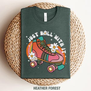 May include: A heather forest green t-shirt featuring a graphic of a pink roller skate with yellow laces, surrounded by white daisies and a colorful rainbow arch. The text "JUST ROLL WITH IT" is printed in a white arched font above the skate.